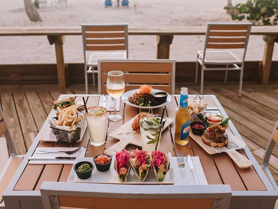 dining-img1 - Cape Eleuthera Resort And Marina, Eleuthera Bahamas Resorts Beachside wooden table with a spread: tacos, fries, burger, and drinks on a decking area by the sand? (informative)