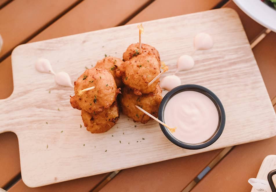 dining-img2 - Cape Eleuthera Resort And Marina, Eleuthera Bahamas Resorts Pile of golden fried croquettes with skewers and a creamy dipping sauce on a light wooden board.