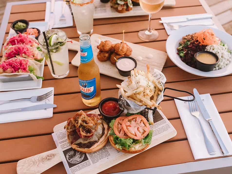 dining-img3 - Cape Eleuthera Resort And Marina, Eleuthera Bahamas Resorts Outdoor wooden table meal: burgers with fries, tomato, greens, dipping sauce, beer bottle, and two drinks in the background.