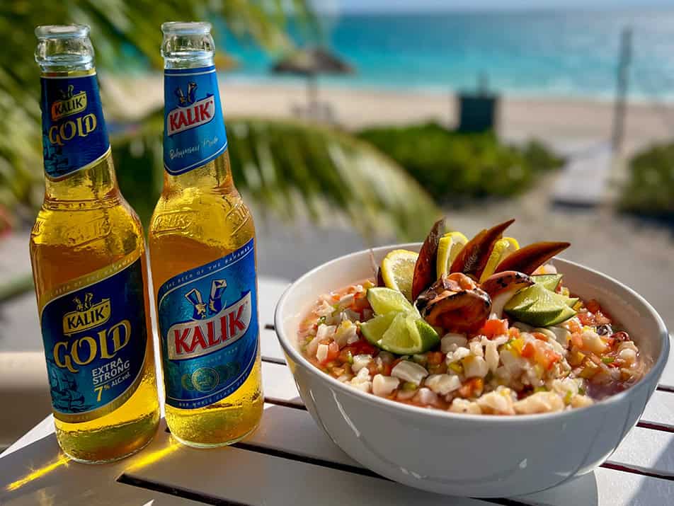 dining-img5 - Cape Eleuthera Resort And Marina, Eleuthera Bahamas Resorts Two Kalik beer bottles sit on a table beside a bowl of seafood salad with lime wedges, ocean view in the background.