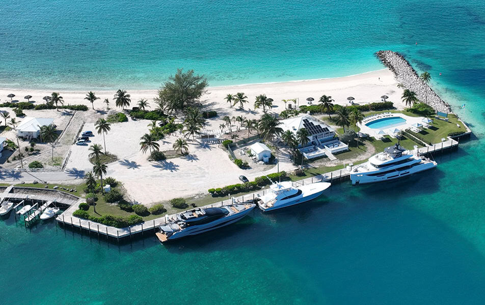 Aerial view of a small tropical island resort with a white-sand beach, palm trees, and several yachts docked at a marina.