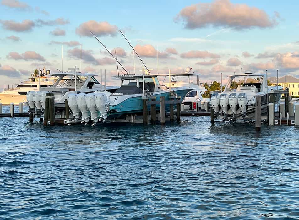 Marina with several motor yachts docked at floating slips and multiple outboard engines visible on the sterns, calm blue water and a partly cloudy sky.