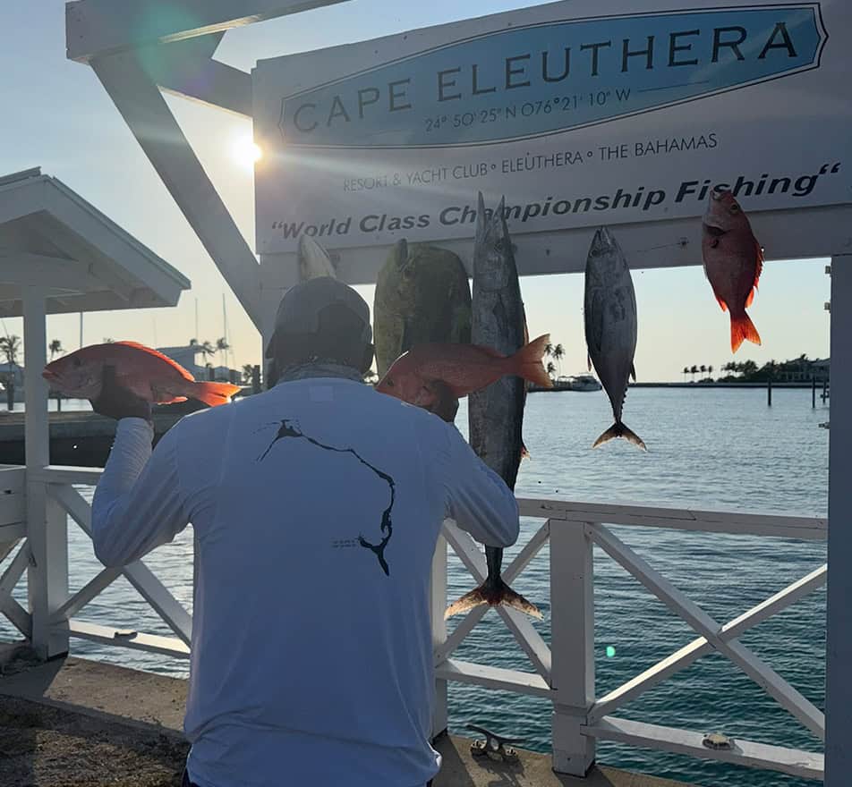 Fisherman in a white long-sleeve shirt stands at a dock with red and gray fish hanging on a display beside a Cape Eleuthera sign at sunset.