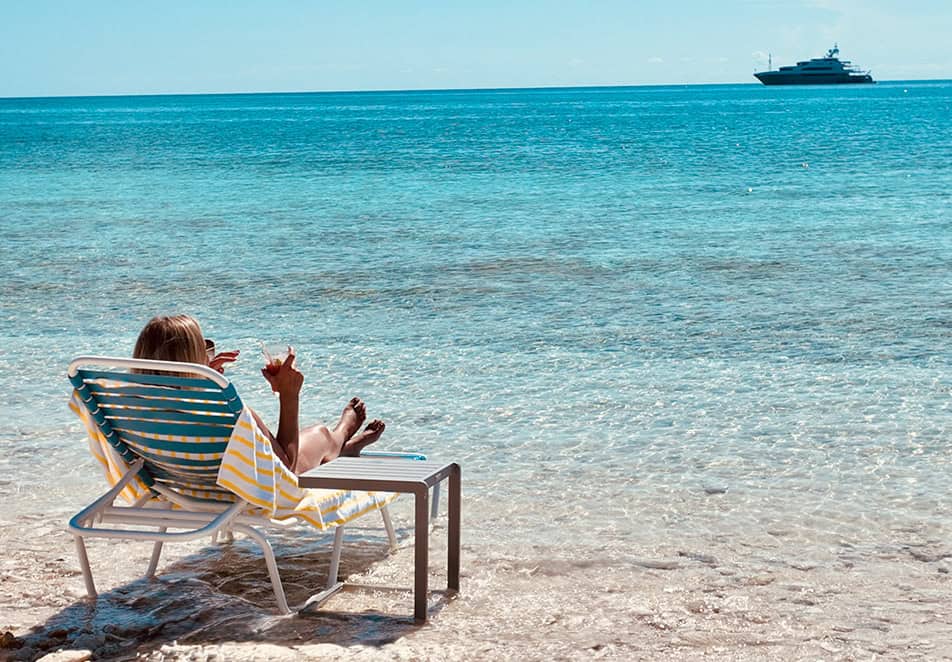 Person relaxing on a striped beach chair by clear turquoise water with a distant yacht on the horizon.