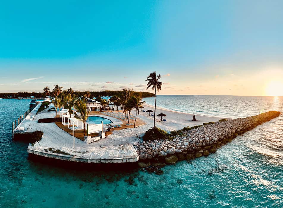 Palm-lined tropical island resort with a pool, gazebo, and a wooden pier, surrounded by turquoise water at sunset.